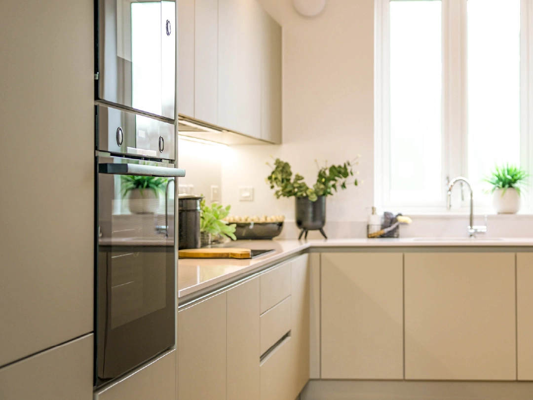 Modern kitchen with sleek cabinets, built-in oven, fresh plants, and natural light from large windows. Minimalist and inviti