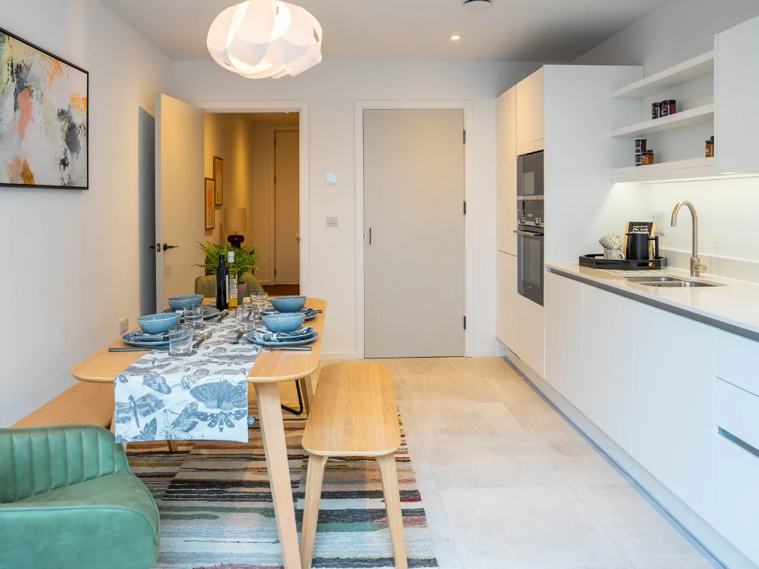 A modern white kitchen with a neutral tiled floor, wooden pre-set dining table with benches and a green chair.