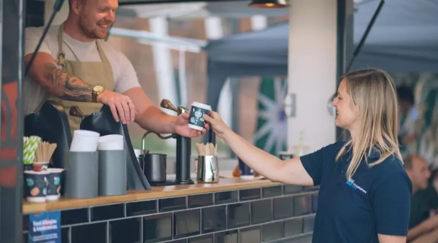 Barista smiles while handing take away coffee to customer outside a pop-up coffee cart at Brabazon in Bristol.