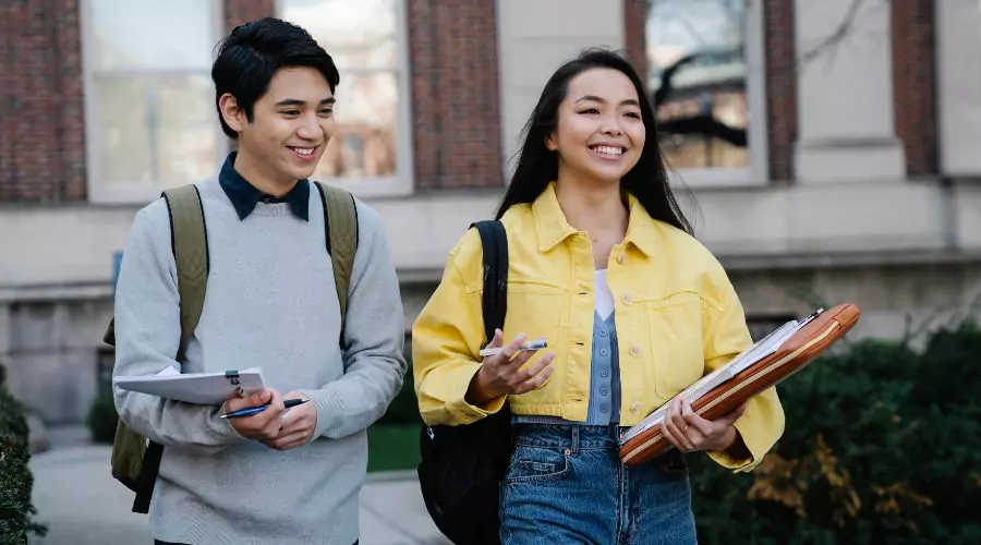 Two students walk together on campus, one holding university papers and the other carrying a laptop and a pen.