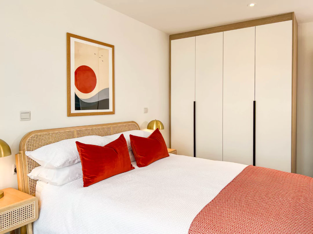 Modern and stylish primary bedroom with a wicker-framed double bed and side tables, white linen duvet with red pillows and white wardrobes. 