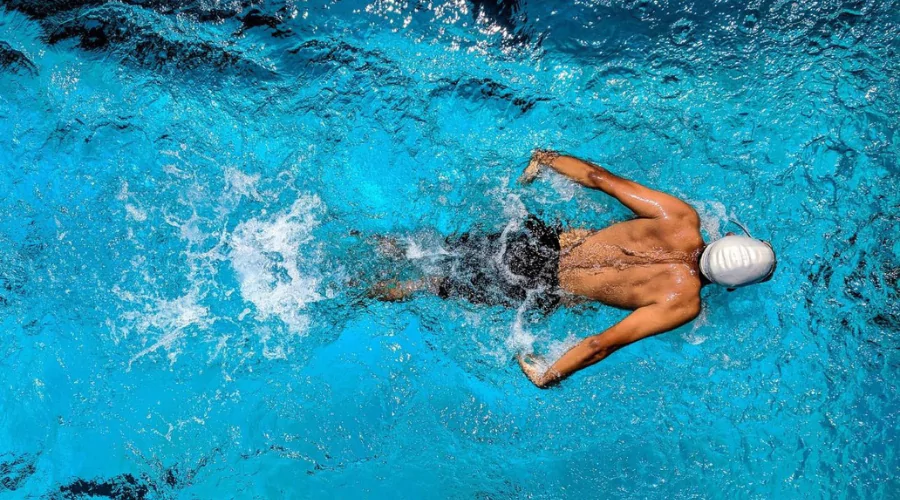 Aerial shot of male wearing black swimming shorts and a white swimming cap training in a swimming pool doing butterfly stroke. 
